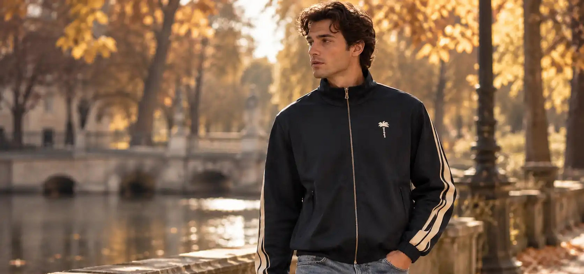 A man wearing a black zip-up track jacket with white stripes on the sleeves and a small white embroidered logo on the chest, standing outdoors by a river with autumn trees in the background.