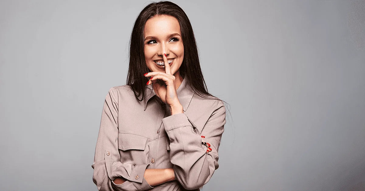 Woman in a beige jacket posing with a finger on her lips against a gray background