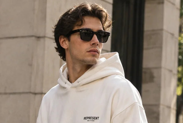 A man wearing a cream-colored hoodie with a black 'REPRESENT' logo on the chest and black sunglasses, standing outdoors against a stone building.