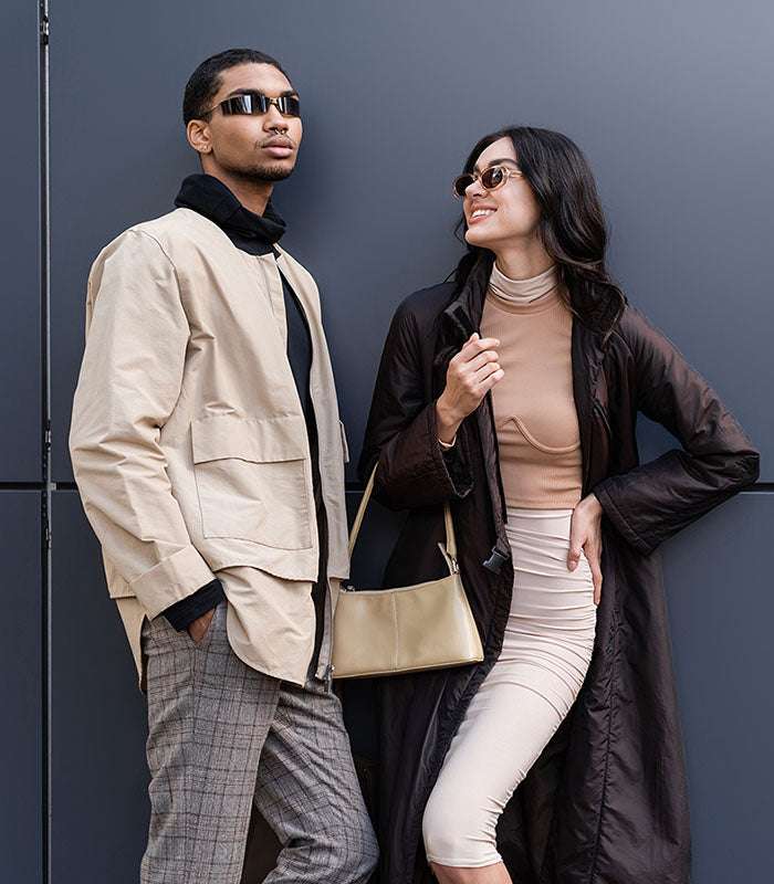 Two people standing against a dark wall, one in a beige coat and sunglasses, the other in a black coat and sunglasses.